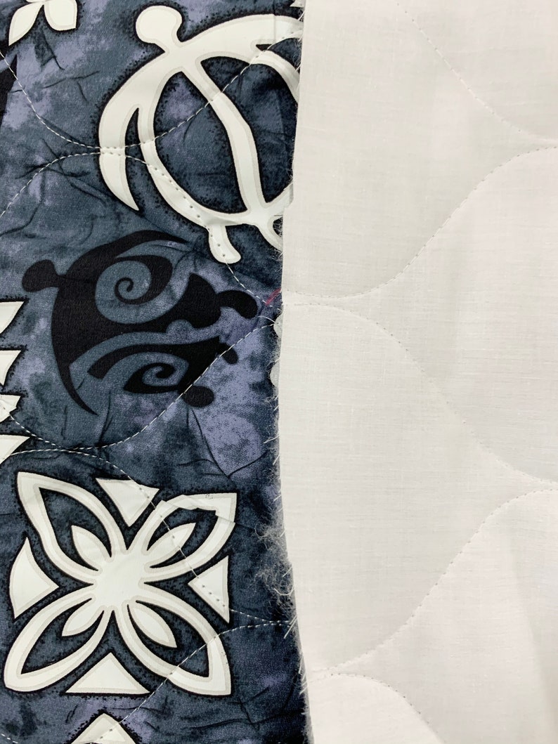 Op de afbeelding: Een close-up van een grijze en witte stof met een gestikt patroon. De stof heeft een herhalend ontwerp van gestileerde bloemen en schildpadden. De stof is genaaid aan een witte stof met een gestikt patroon.