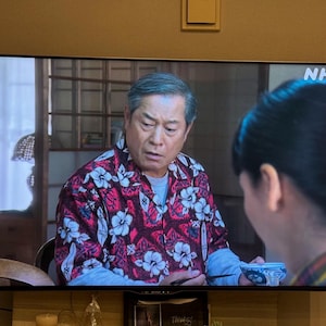 Könnte beinhalten: Ein Mann in einem rot-weiß gemusterten Hemd sitzt auf einem Stuhl. Er blickt mit ernster Miene in die Kamera. Der Hintergrund ist ein verschwommenes Bild eines Raumes mit einem Fenster. Der Text "NHK" ist in der oberen rechten Ecke des Bildes sichtbar.