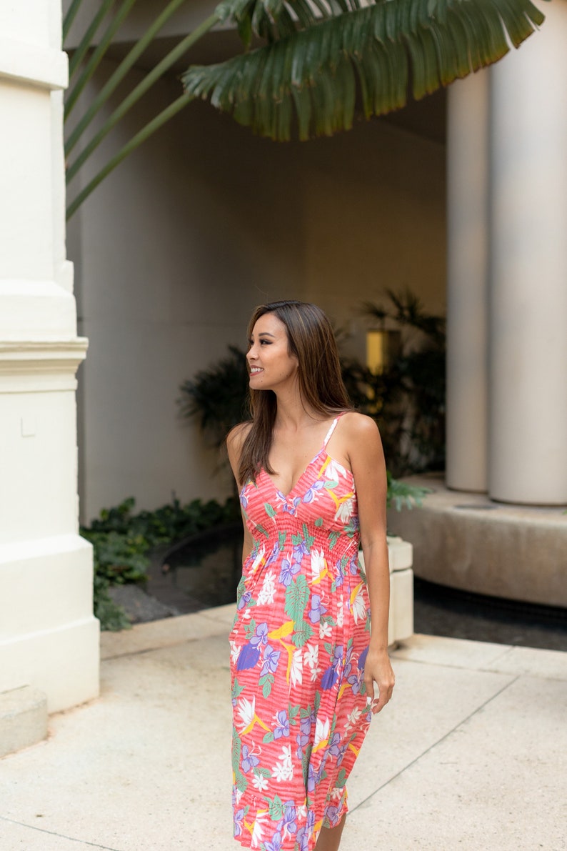 May include: A woman in a pink floral midi dress with a V-neckline and thin straps. She is standing in a courtyard with a white wall and green plants in the background.