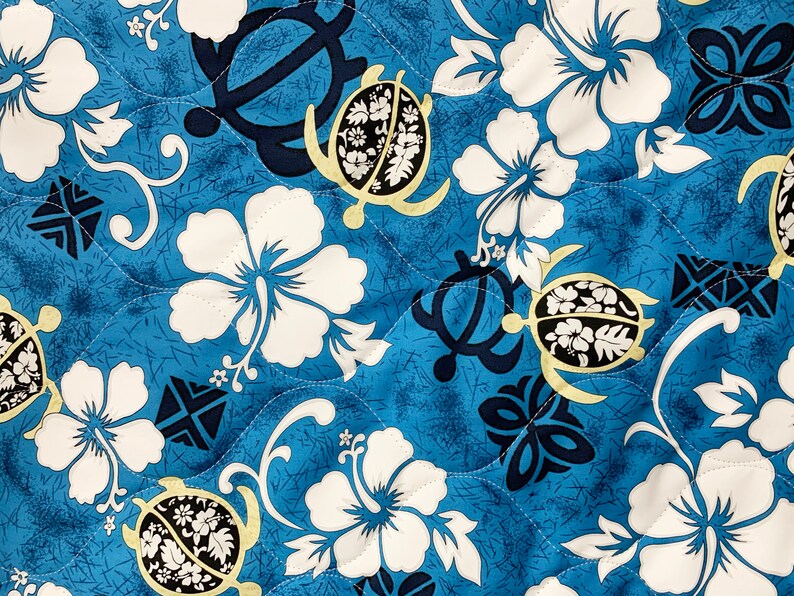 Op de afbeelding: Blauwe stof met een herhalend patroon van witte hibiscusbloemen, gele en zwarte zeeschildpadden en donkerblauwe geometrische vormen.