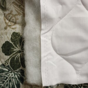 May include: Close-up of a white quilted fabric with a diamond pattern. The fabric is sewn to a beige fabric with a green floral pattern.