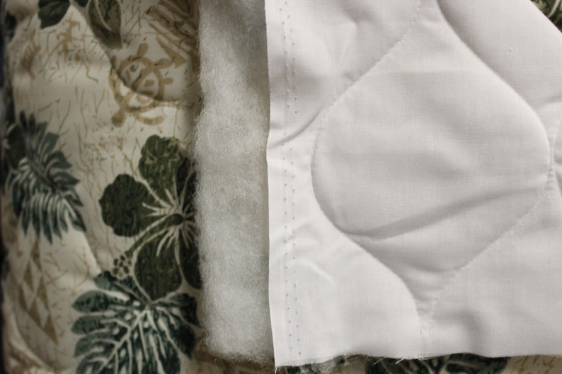 May include: Close-up of a white quilted fabric with a diamond pattern. The fabric is sewn to a beige fabric with a green floral pattern.