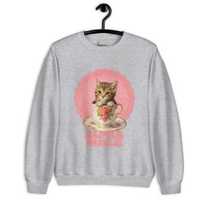 Könnte beinhalten: Ein graumeliertes Sweatshirt mit Rundhalsausschnitt und einem Grafikdruck eines braunen getigerten Kätzchens, das in einer geblümten Teetasse sitzt. Das Kätzchen ist auf einem rosa, gebogten Spitzenhintergrund zentriert. Darunter steht ALPHA MALE.