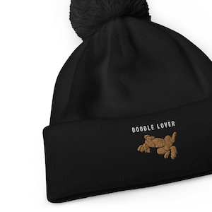 Puede incluir: Gorro negro con un pompón esponjoso en la parte superior. El gorro tiene un perro doodle bordado en blanco y el texto "Doodle Lover" en blanco.