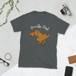 May include: A dark heather grey t-shirt with a doodle-dad design. The design features a brown doodle dog sleeping on its back with its paws in the air. The text "Doodle-Dad" is printed in white above the dog.