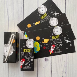 Puede incluir: Caja de jugo negra y blanca con un gráfico de cohete y luna. El texto "ROCKET FUEL 2 the moon" está impreso en la caja.