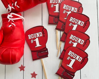 May include: Red boxing gloves and several red boxing glove-shaped cupcake toppers with the text "ROUND #1" in white. The toppers are on wooden sticks. Red star-shaped confetti is scattered around the items.