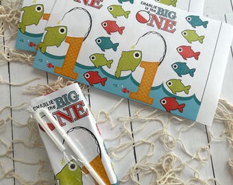May include: Party favor bags with a fishing theme. The bags are white with the text "Charlie is the BIG ONE" and feature illustrations of fish, fishing poles, and waves. A juice box with straws is also visible.