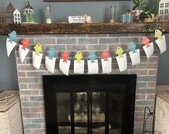 May include: A decorative banner featuring colorful fish-shaped clips holding white paper tags. The fish are in shades of blue, red, and yellow, with the text "MONTHLY FIVE" printed on the tags. The banner is strung across a brick fireplace.