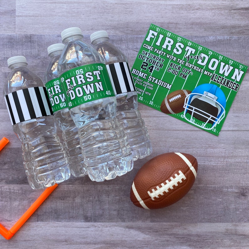 May include: Four clear plastic water bottles with black and white striped labels that say "First Down" and green and white labels with football yard lines. A brown football and an orange plastic football goal post are on a wooden surface. A green and white invitation with the text "First Down" and "Come party with the birthday MVP Alexander" is in the background.