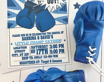 May include: Blue boxing gloves and a baby shower invitation with the text "This baby is going to be... A Knock Out!" The invitation includes event details and star-shaped confetti.