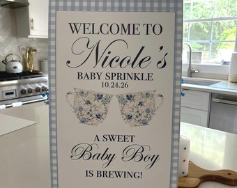 May include: A rectangular sign with a light blue gingham border featuring the text WELCOME TO Nicole's BABY SPRINKLE 10.24.26, two floral teacup illustrations, and the text A SWEET Baby Boy IS BREWING! in dark blue and black lettering.