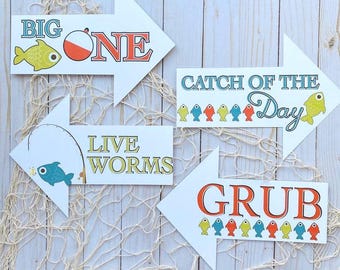 May include: Four white arrow-shaped signs with fishing-themed text and illustrations. The signs read "BIG ONE", "LIVE WORMS", "CATCH OF THE Day", and "GRUB". Each sign has a different fish illustration and is set against a wooden background.