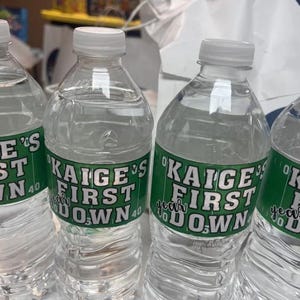 May include: Three clear plastic water bottles with green and white labels that say "Kaige's First Down" with a football field graphic.