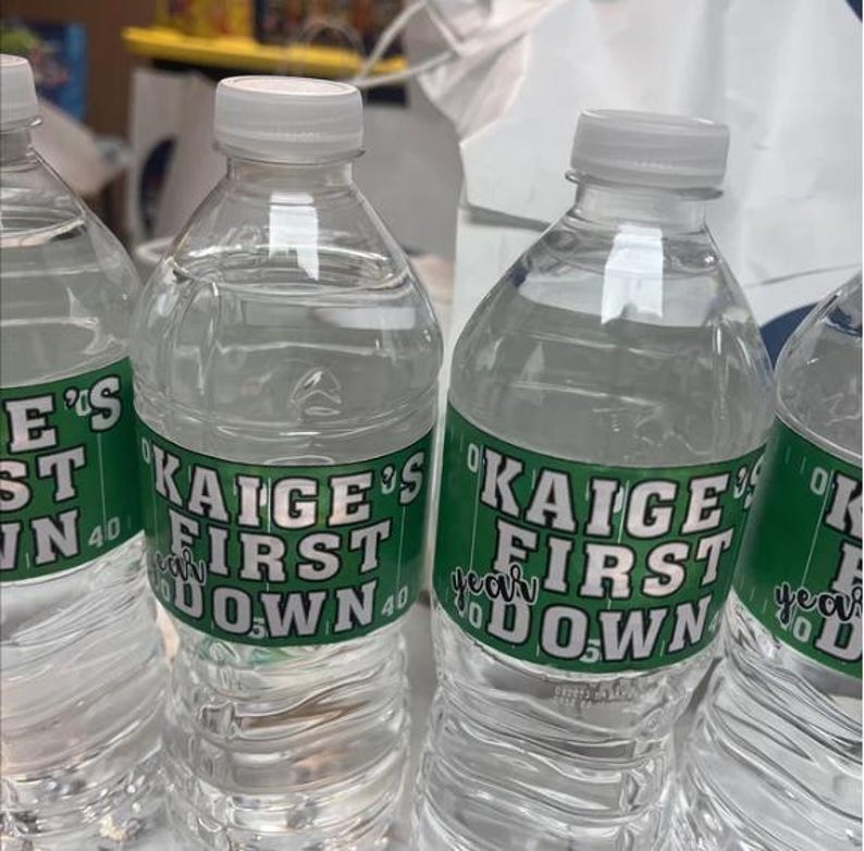 May include: Three clear plastic water bottles with green and white labels that say "Kaige's First Down" with a football field graphic.