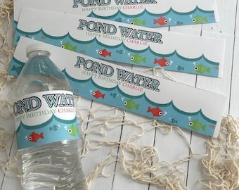 May include: A clear plastic water bottle with a label that reads "POND WATER HAPPY BIRTHDAY CHARLIE". The label has a blue wave design with red, green, and blue fish. Several matching labels are also visible.