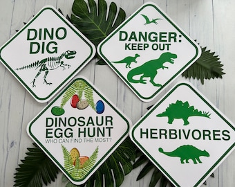 May include: Four white diamond-shaped signs with green borders and dinosaur-themed text and illustrations. The signs read "Dino Dig", "Danger: Keep Out", "Dinosaur Egg Hunt: Who Can Find The Most?", and "Herbivores".