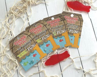 May include: A collection of fishing-themed party favor tags and red fish-shaped candies. The tags read "Thank You for making my party REEL FUN!" and feature fish and the number "1". The background includes a fishing net.