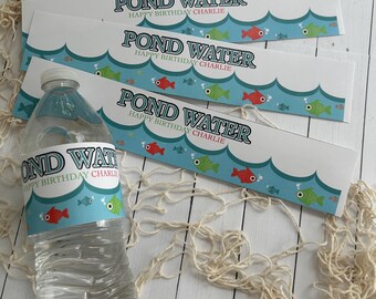May include: A clear plastic water bottle with a label that reads "POND WATER HAPPY BIRTHDAY CHARLIE" is displayed with matching decorative paper bands. The bands and bottle label feature a blue wave design with red, green, and blue fish illustrations.