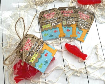 May include: A collection of party favors featuring red fish-shaped candies and tags. The tags have a wooden plank design with the text "THANK YOU for making my party REEL FUN!" and a number one. The favors are arranged on a white wooden surface with a fishing net.