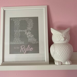 Op de afbeelding: Een ingelijst afdruk met een grijze achtergrond en witte tekst. De tekst is een gedicht over een babymeisje genaamd Rylie. Het gedicht is geschreven in een lettertype dat eruitziet als een handgeschreven brief. De lijst is wit en heeft een witte passe-partout. De afdruk staat op een wit rek met een wit keramiek uilenbeeld rechts van de afdruk.