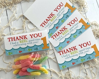 May include: A collection of party favors featuring a fishing theme. The set includes thank you cards with a fish and wave design, the number 1, and a bag of colorful gummy candies. The text on the cards reads "Thank You, I had a 'REEL' good time, celebrating with you!"