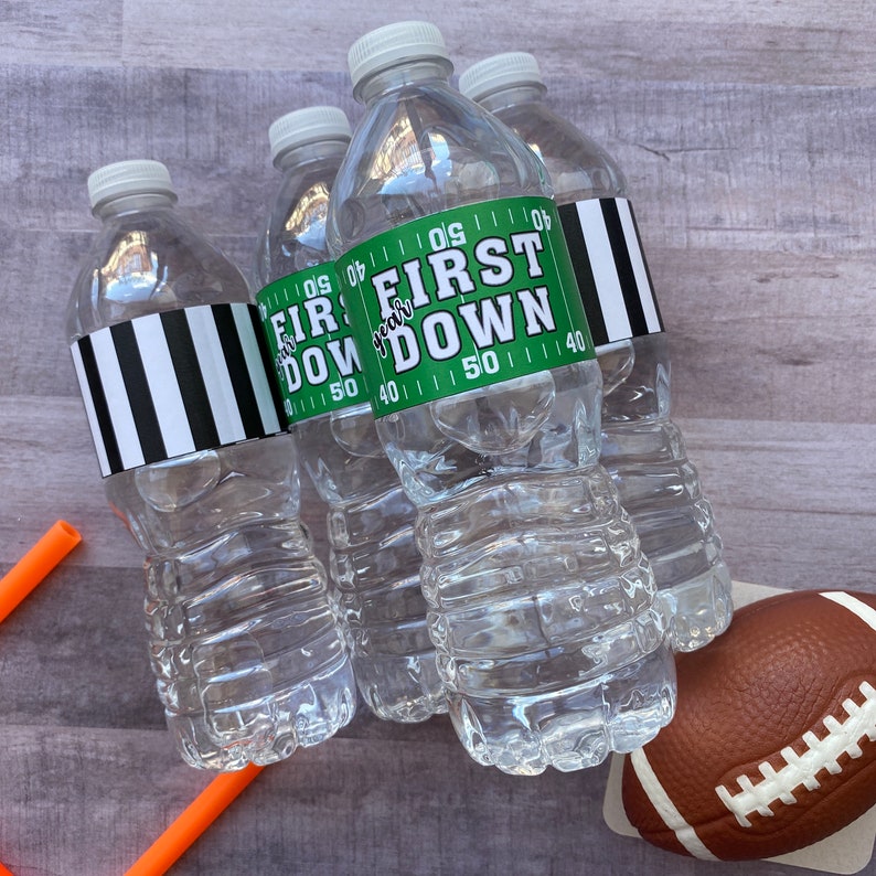 May include: Four clear plastic water bottles with green and white labels that say "First Down" with a football field graphic. The labels also have black and white stripes. A brown football is in the bottom right corner of the image.