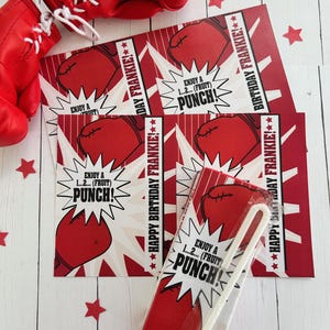 May include: Red boxing gloves and birthday-themed juice boxes with the text "Enjoy a 1...2... (Fruit) Punch!" and "Happy Birthday Frankie!" on a white wooden surface. Red star-shaped confetti is scattered around.
