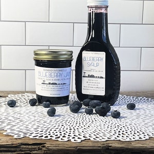 May include: Two glass jars with labels, one filled with blueberry jam and the other with blueberry syrup. Both jars have the same brand name, "Soondook Enterprises LLC". The jars are sitting on a white doily with blueberries scattered around them.