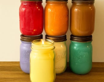 May include: A collection of seven glass mason jar candles with dark metal lids. The candles feature wax in various colors including red, orange, brown, purple, yellow, white, and teal. One yellow candle in the front is lit with a visible flame.