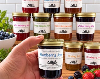 Peut inclure: Une collection de bocaux en verre remplis de diverses confitures et gelées, chacun doté d'un couvercle doré et d'une étiquette blanche avec le logo Boondock Enterprises LLC. Les étiquettes indiquent les ingrédients et des parfums variés.