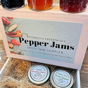 May include: A white box with the text "Boondock Enterprises Pepper Jams Spicy Jam Sampler" on the front. The box contains six 4-ounce jars of spicy pepper flavored jams or jellies. The jars are labeled with the flavors "Strawberry Jalapeno", "Sweet Jalapeno", and "Jalapeno Flavor".