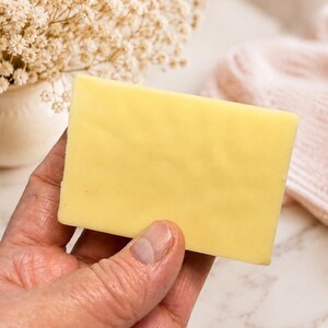 May include: A rectangular bar of yellow soap is held in a hand. The soap has a smooth surface and a pale yellow color. The background includes a vase of white flowers and a pink knitted item.