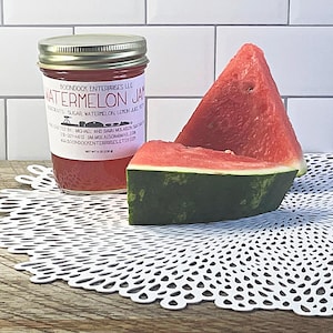 Puede incluir: Un frasco de vidrio de mermelada de sand&iacute;a con tapa plateada y etiqueta blanca que dice Watermelon Jam, Boondock Enterprises LLC, y Net Wt 8 oz (226 g), acompa&ntilde;ado de dos rodajas triangulares de sand&iacute;a roja con corteza verde.