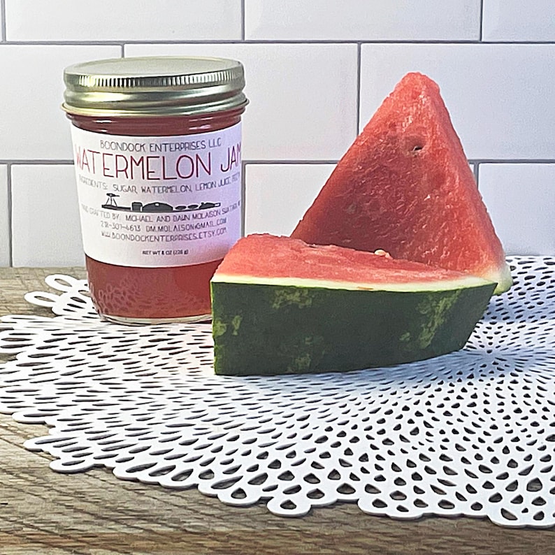 Puede incluir: Un frasco de vidrio de mermelada de sand&iacute;a con tapa plateada y etiqueta blanca que dice Watermelon Jam, Boondock Enterprises LLC, y Net Wt 8 oz (226 g), acompa&ntilde;ado de dos rodajas triangulares de sand&iacute;a roja con corteza verde.
