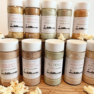 May include: Ten glass jars with white lids filled with popcorn seasoning. The jars are arranged in two rows of five on a wooden surface. The jars are labeled with different flavors, including Dorito, Smokey BBQ, Ranch, Parmesan Garlic, Taco, Cream Cheese Cinnamon Roll, Chocolate Peanut Butter, Dill Pickle, Salted Caramel, and Jalapeno White Cheddar.