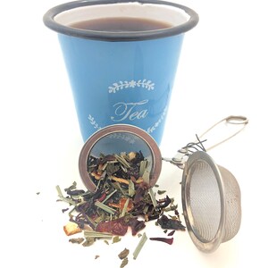 May include: A blue enamel mug with the word "Tea" printed on it, filled with dark tea. A metal tea infuser with loose leaf tea is next to the mug.