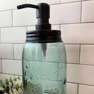 May include: A glass soap dispenser with a dark brown metal pump and lid. The dispenser is filled with clear liquid and has a textured glass design.