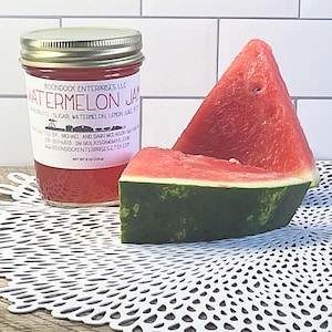Puede incluir: Un frasco de vidrio de mermelada de sand&iacute;a con tapa plateada y etiqueta blanca que dice Watermelon Jam, Boondock Enterprises LLC y Net Wt 8 oz (226 g), acompa&ntilde;ado de dos rebanadas triangulares de sand&iacute;a roja con corteza verde.