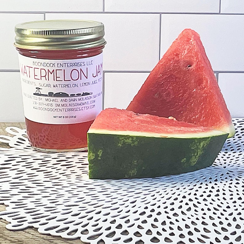 Puede incluir: Un frasco de vidrio de mermelada de sand&iacute;a con tapa plateada y etiqueta blanca que dice Watermelon Jam, Boondock Enterprises LLC y Net Wt 8 oz (226 g), acompa&ntilde;ado de dos rebanadas triangulares de sand&iacute;a roja con corteza verde.