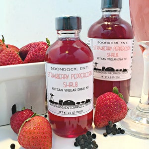 Op de afbeelding: Twee glazen flessen met aardbei-peperkornsap, een rode vloeistof erin, met een wit etiket waarop staat "Boondock, Ent. Strawberry Peppercorn Shrub Artisan Vinegar Drink Mix" en een nettogewicht van 4,2 oz (125 g). Op de achtergrond een glas met roze vloeistof en aardbeien.