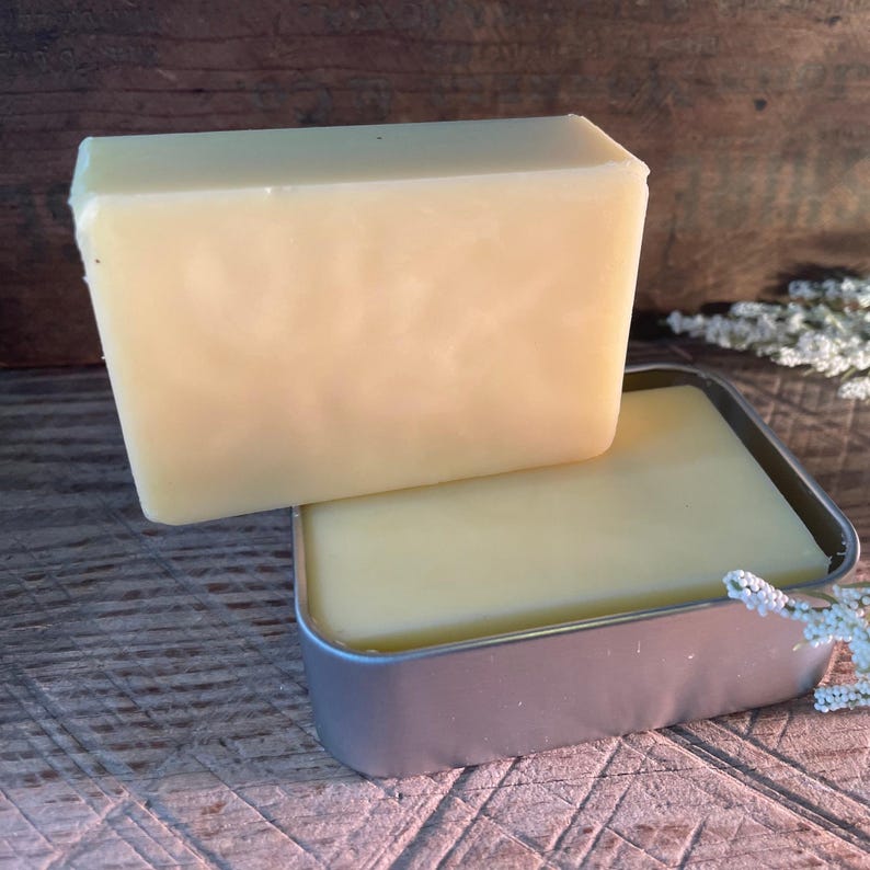 May include: A light yellow bar of soap rests atop a rectangular metal tin containing a similar soap. The soap has a smooth, solid appearance. The background is a dark wooden surface.