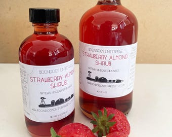 Op de afbeelding: Twee glazen flessen aardbeien-amandelsiroop met zwarte doppen en witte etiketten. De etiketten luiden "Strawberry Almond Shrub" en "Artisan Vinegar Drink Mixer". Verse aardbeien staan op de voorgrond. Eén fles is 118 ml, de andere 237 ml.