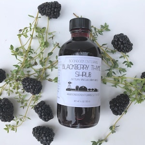 Op de afbeelding: Een glazen fles met een zwart etiket waarop "BOONDOCK ENTERPRISE BLACKBERRY THME SHRUB ARTISAN VINEGAR DRINK MIXER WABCONDOCHENTERREESETS.COM NET WT 4 OZ (113G)" staat. De fles is omringd door bramen en tijmtakjes op een witte achtergrond.