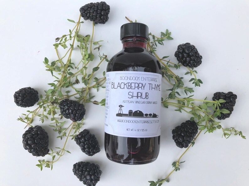 Op de afbeelding: Een glazen fles met een zwart etiket waarop "BOONDOCK ENTERPRISE BLACKBERRY THME SHRUB ARTISAN VINEGAR DRINK MIXER WABCONDOCHENTERREESETS.COM NET WT 4 OZ (113G)" staat. De fles is omringd door bramen en tijmtakjes op een witte achtergrond.