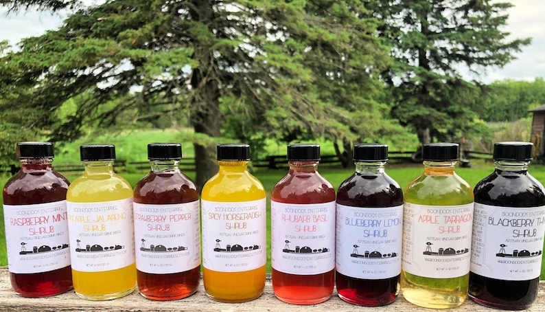 Op de afbeelding: Acht glazen flessen met verschillende gekleurde siropen met etiketten waarop staat "Raspberry Mint Shrub", "Earl Jalapeno Shrub", "Strawberry Pepper Shrub", "Spicy Horseradish Shrub", "Rhubarb Basil Shrub", "Blueberry Lemon Shrub", "Apple Tarragon Shrub" en "Blackberry Thyme Shrub".