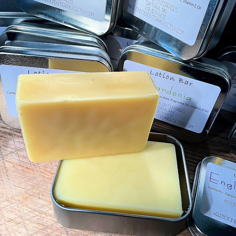 May include: A close-up of two yellow lotion bars, one resting on top of the other, inside small, silver tins. The top bar is rectangular. The tins have labels with text, including "Gardenia" and a website address. The bars appear to be handmade.