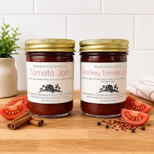 May include: Two glass jars with gold lids containing Tomato Jam and Smokey Tomato Jam. Each label lists ingredients, Boondock Enterprises LLC, and a net weight of 8 oz (226 g). The jars are placed on a wooden surface with tomato slices and cinnamon sticks.