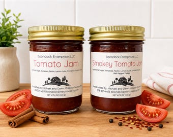 May include: Two glass jars with gold lids containing Tomato Jam and Smokey Tomato Jam. Each label lists ingredients, Boondock Enterprises LLC, and a net weight of 8 oz (226 g). The jars are placed on a wooden surface with tomato slices and cinnamon sticks.