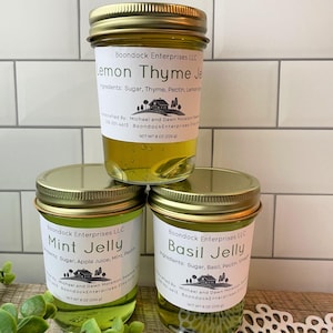 May include: Three glass jars of homemade jelly, stacked on top of each other. The top jar is labeled "Lemon Thyme Jelly" and contains a light yellow jelly. The middle jar is labeled "Mint Jelly" and contains a green jelly. The bottom jar is labeled "Basil Jelly" and contains a green jelly.